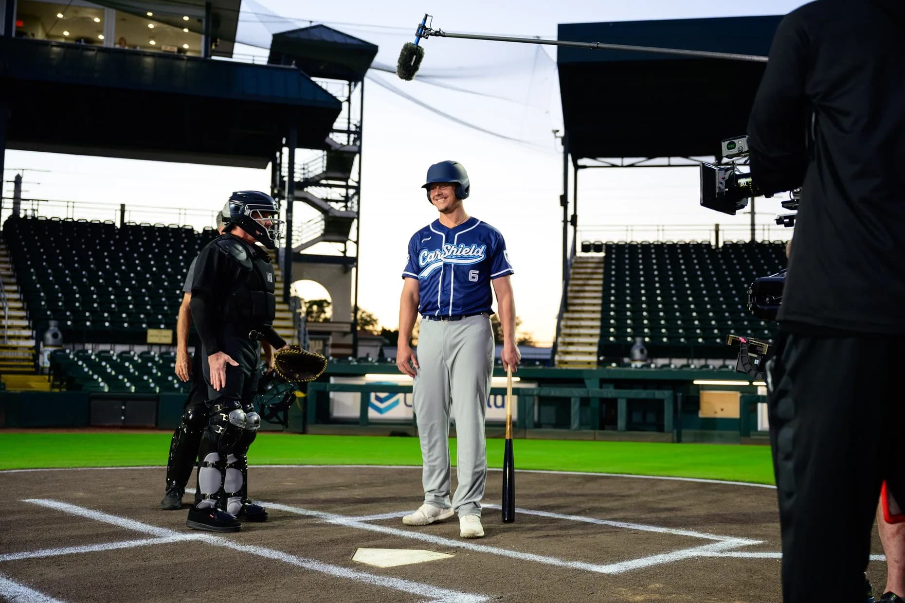 A performer in a CarShield baseball uniform standing at home plate beside a catcher while a camera and boom microphone record