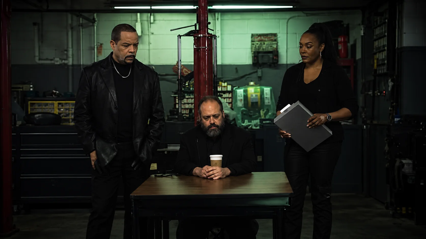 Two performers standing beside a seated man at a table in a dim garage, from a CarShield commercial