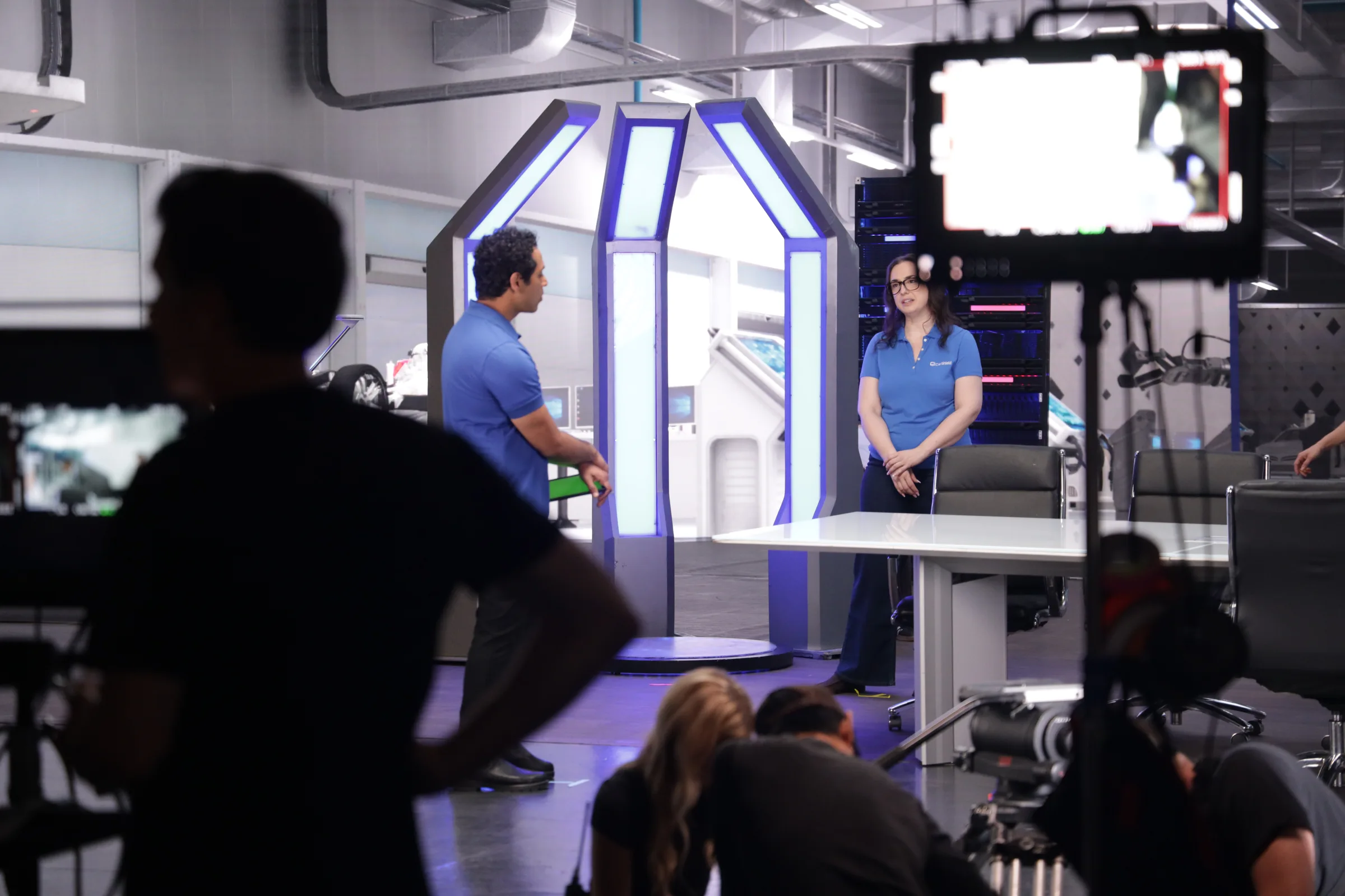 Two performers on a futuristic studio set with crew and a camera monitor in the foreground