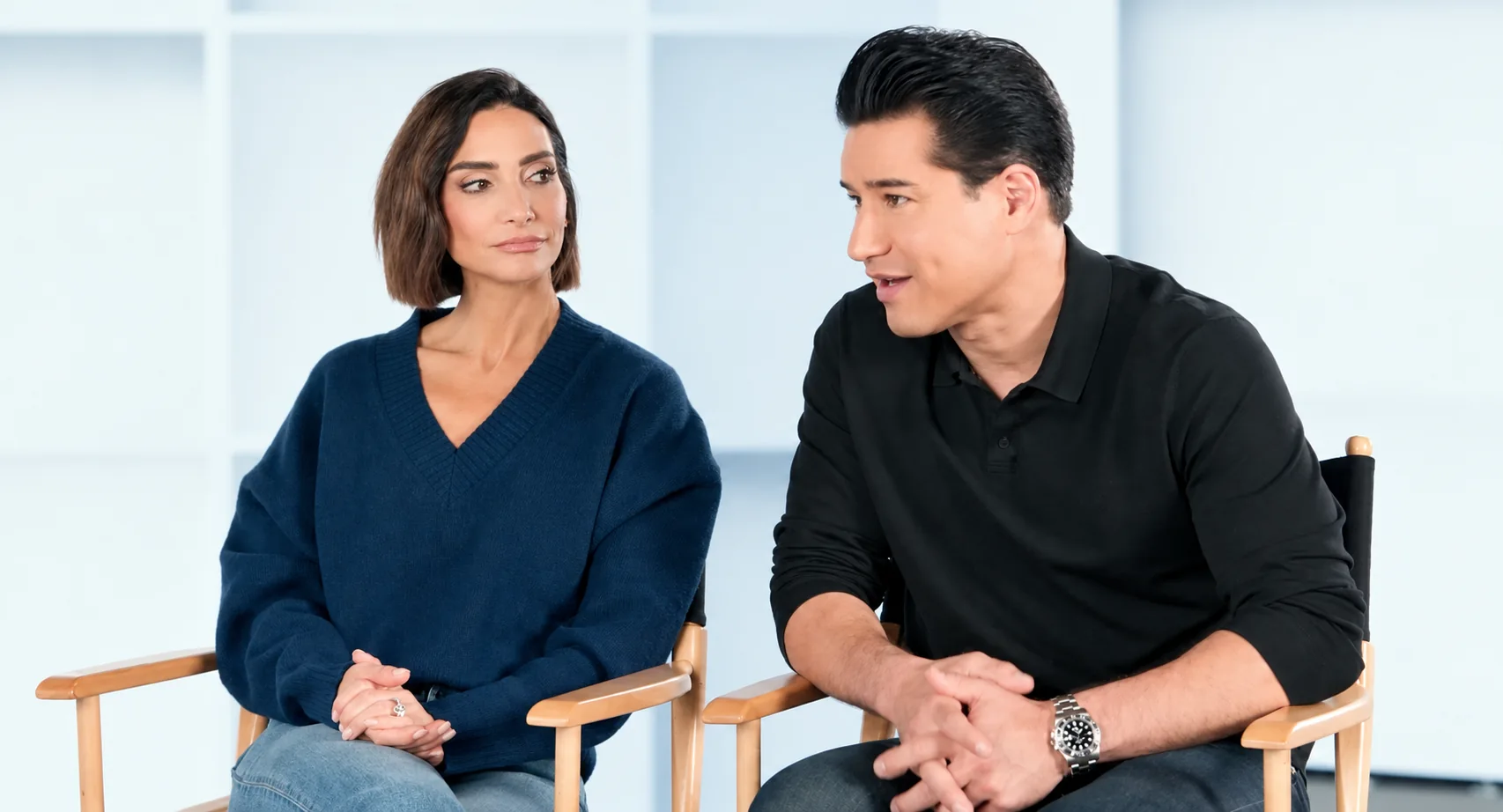A woman in a blue sweater and a man in a black polo seated in director's chairs on a white studio set