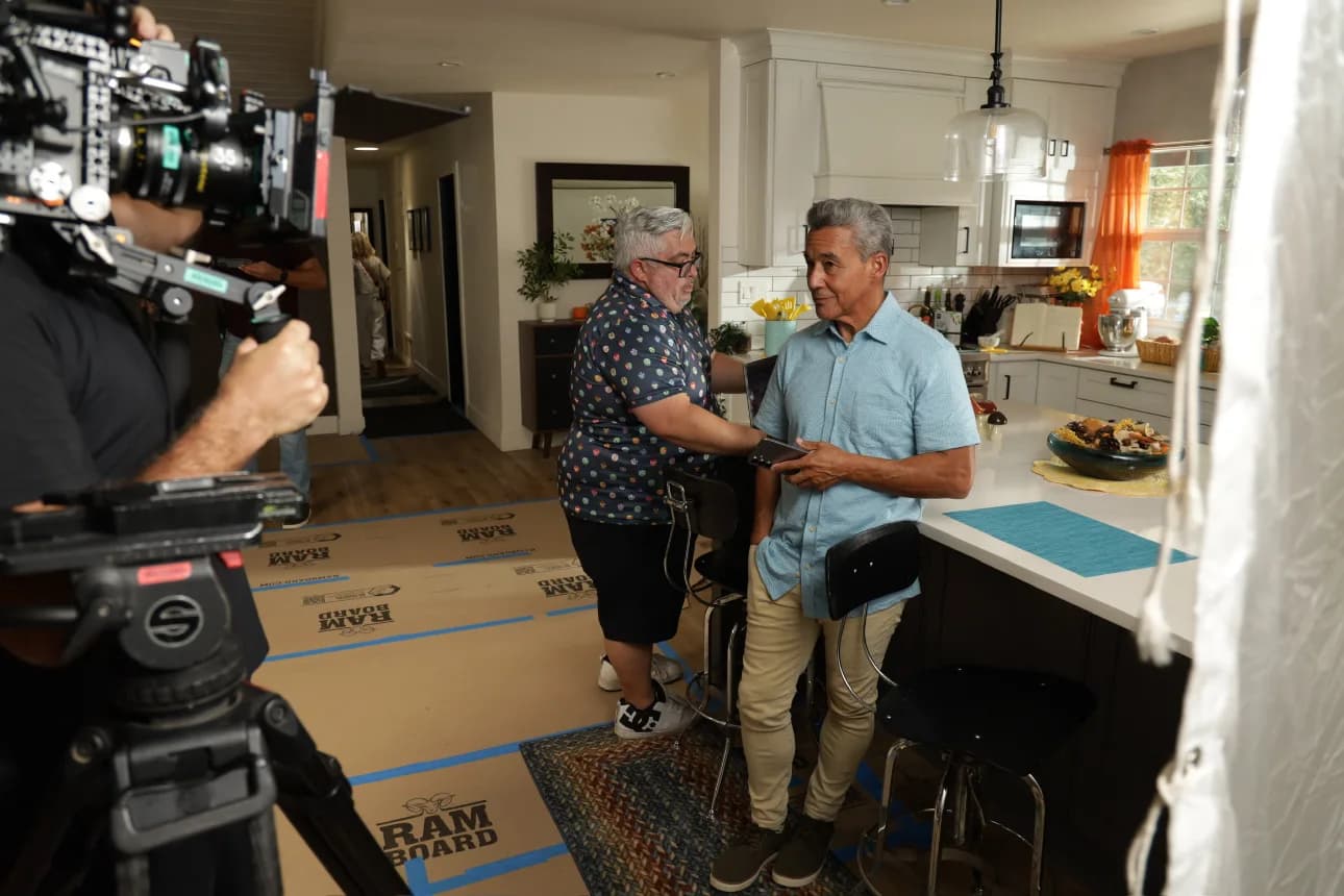 A camera in the foreground films two men talking at a kitchen island on set.
