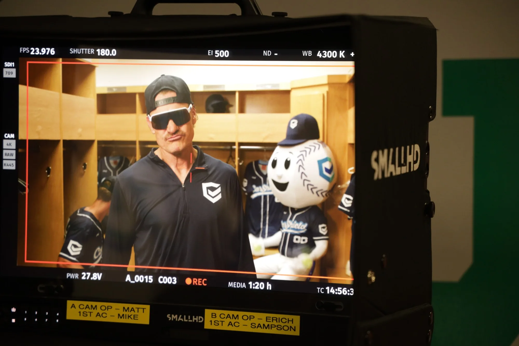 A camera monitor showing a performer in sunglasses beside a baseball mascot in a locker room