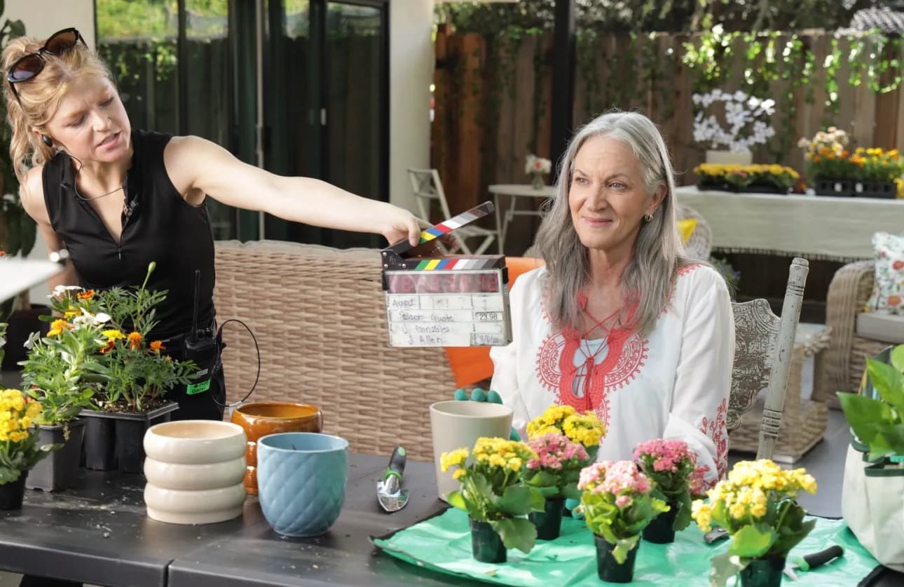 A crew member slates a take beside an actress at a garden potting table on set.